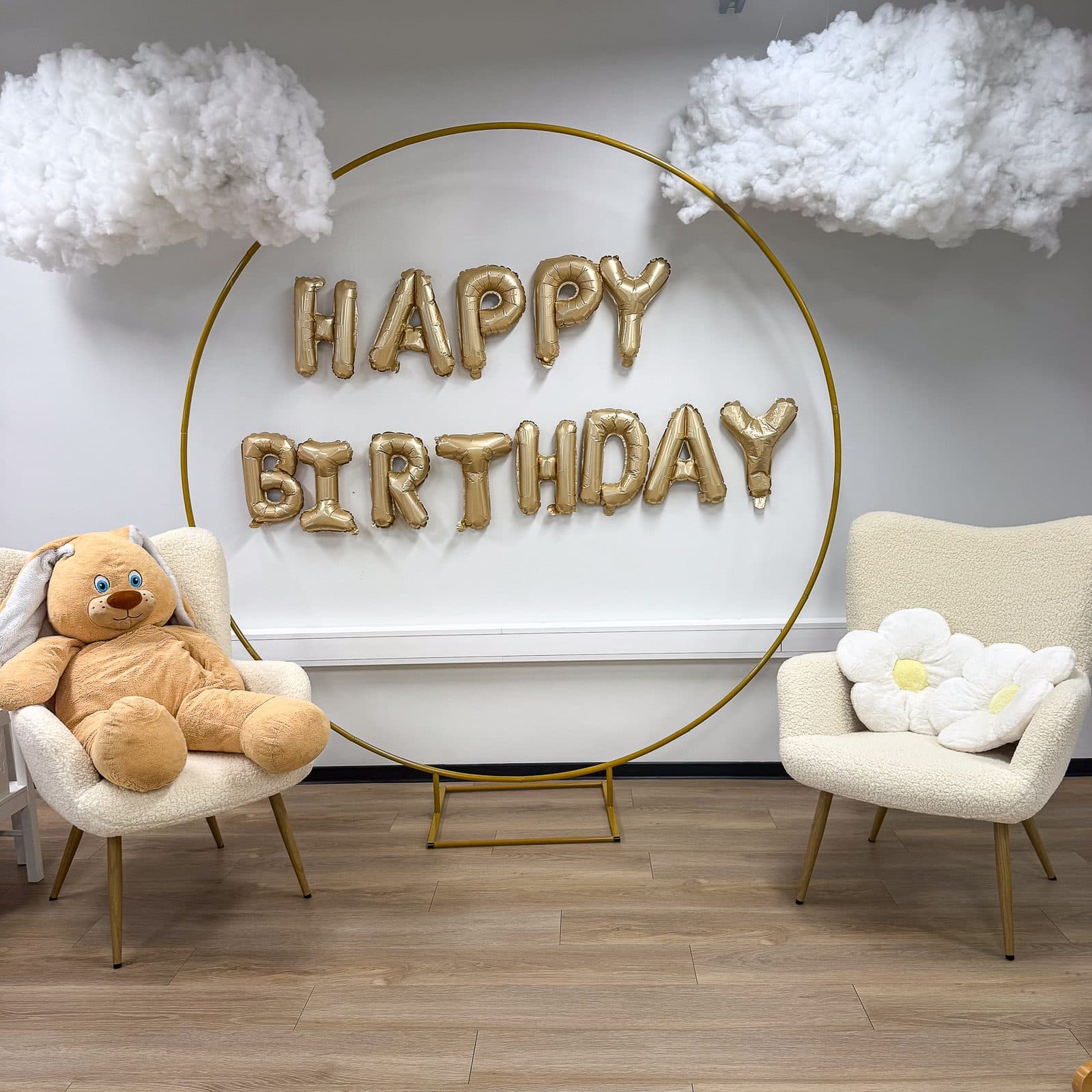 Fotosein: kuldne ringikujuline kaar HAPPY BIRTHDAY õhupallitähtedega, kõrval kaks heledat tugitooli – ühel suur kaisukaru, teisel lillepadi – ja lae all vatipilved
